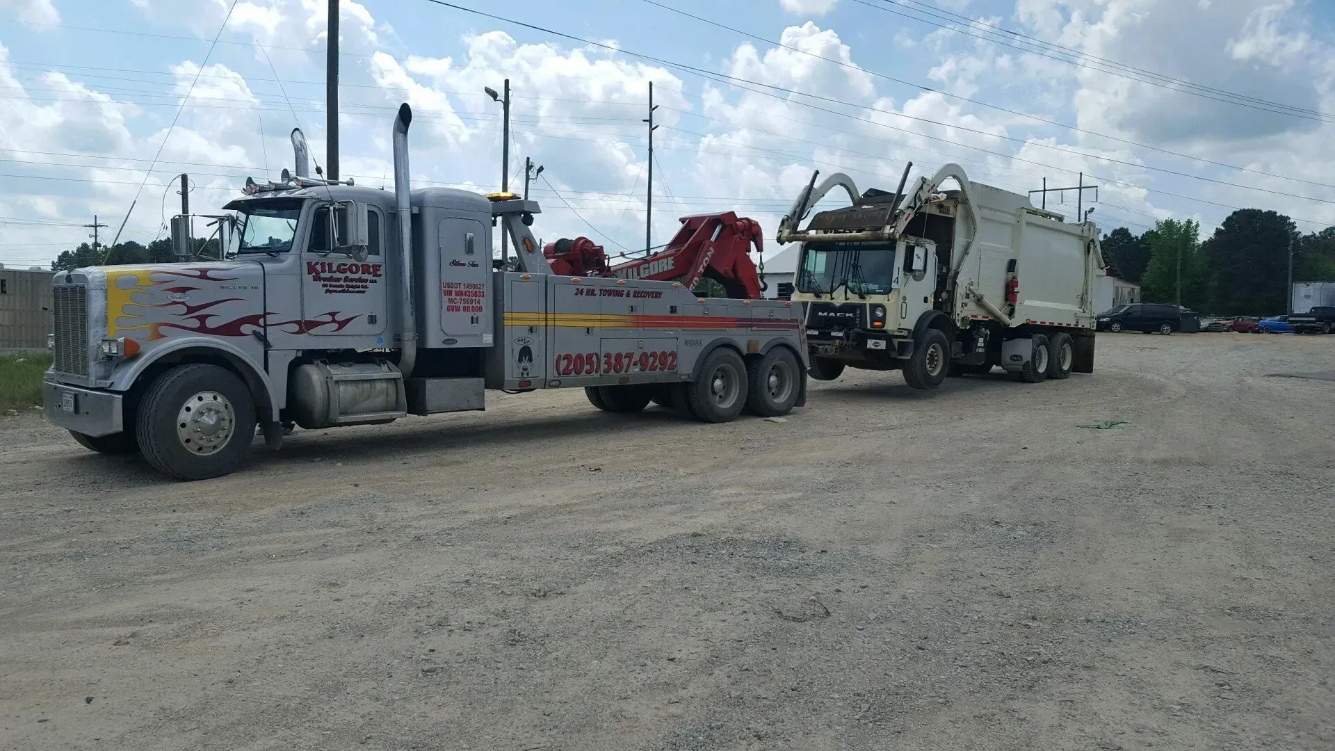 A tow truck is towing a garbage truck in a parking lot.