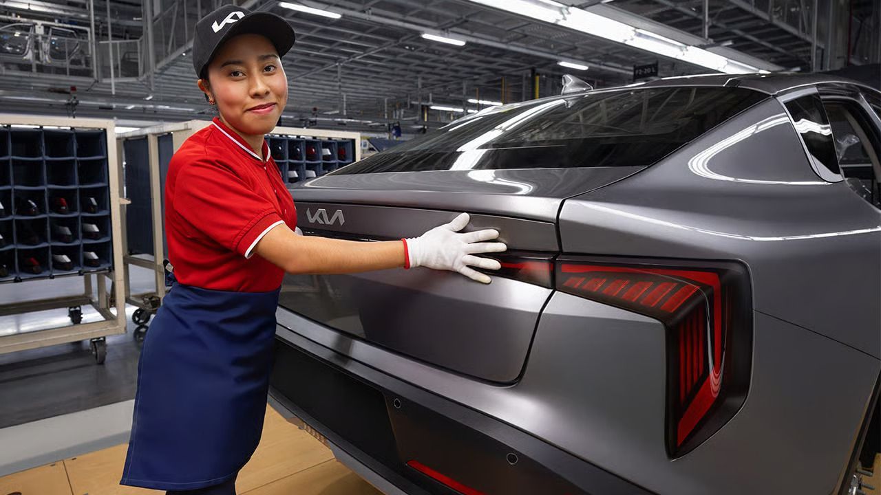Un obrero de fábrica, vestido con una camisa roja y un guante blanco, inspecciona el maletero de un coche kia