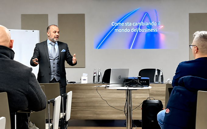 Un relatore tiene una presentazione in una sala conferenze, di fronte a un pubblico, con una diapositiva proiettata alle sue spalle.