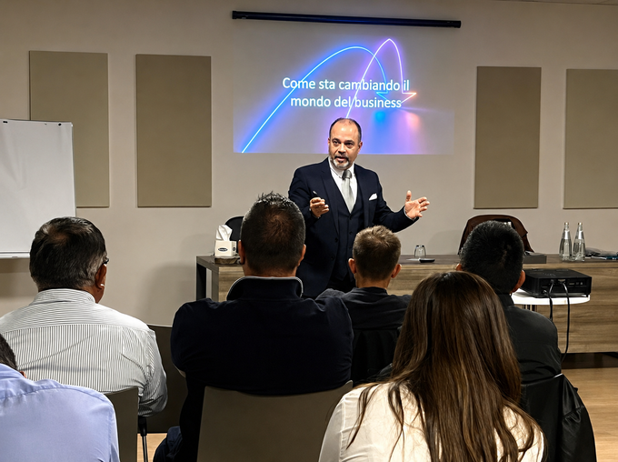 Un relatore tiene una presentazione in una sala conferenze davanti a partecipanti seduti, con una diapositiva proiettata alle sue spalle.