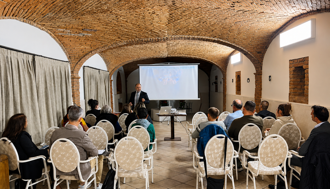 Pubblico seduto in una sala ad arco che assiste a una presentazione proiettata su uno schermo.