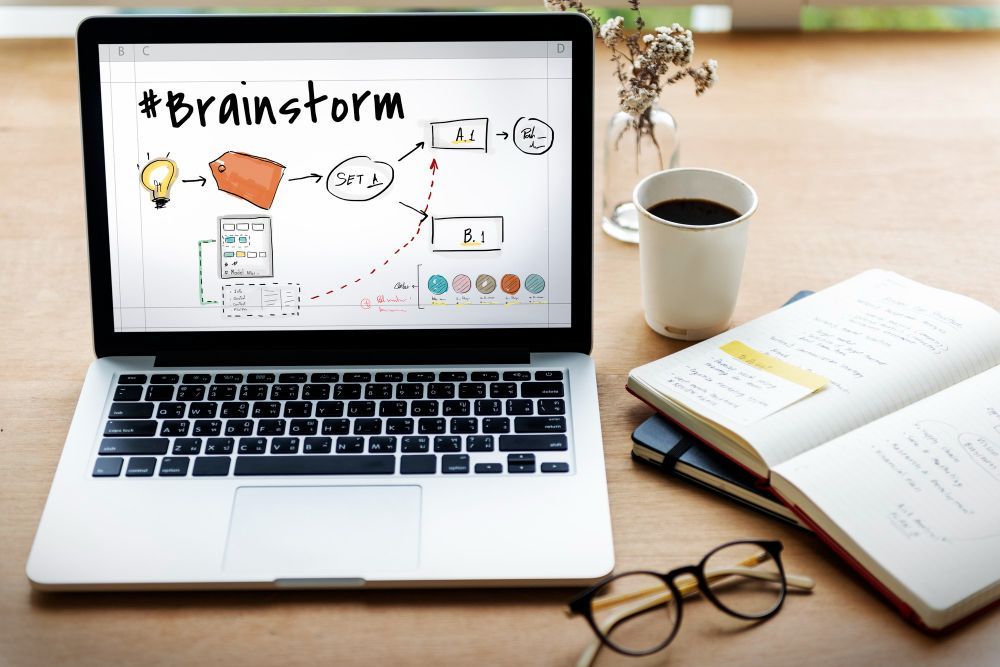 Laptop on desk displaying a brainstorm diagram, beside coffee, notebook, and eyeglasses