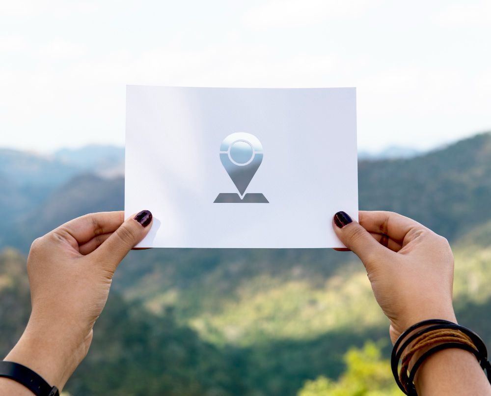Hands holding a white card with a location pin icon against a blurred mountain background
