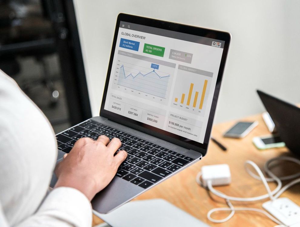 Person using a laptop showing analytics dashboards on a desk with a smartphone and charging cable nearby