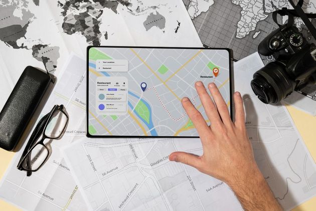 Hand on tablet displaying a map, with camera, glasses, and documents on a desk