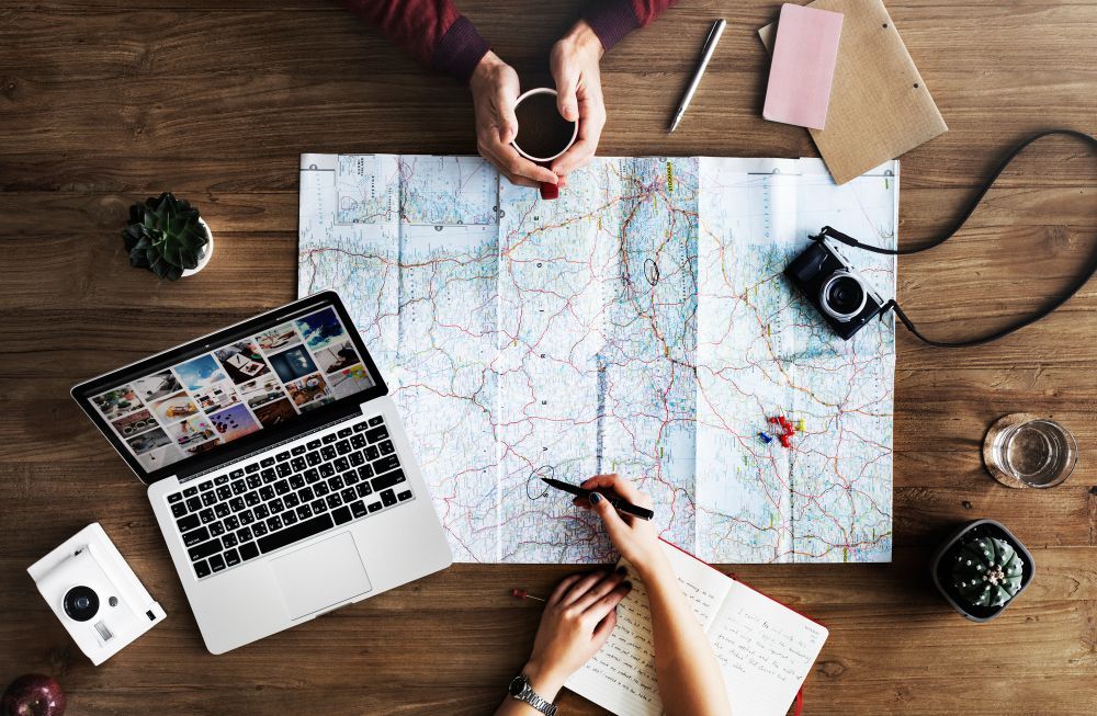 People planning a trip around a large map with notebooks, camera, and laptop on a wooden table
