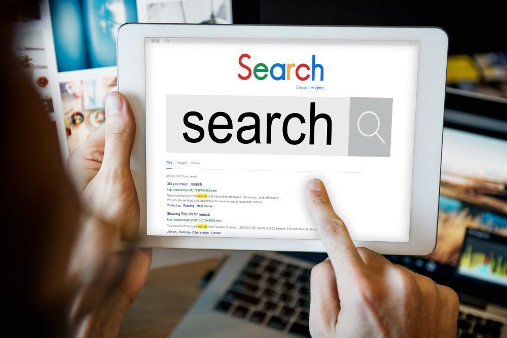 Hands pointing at a tablet showing a search engine page with a large search box and magnifying glass icon