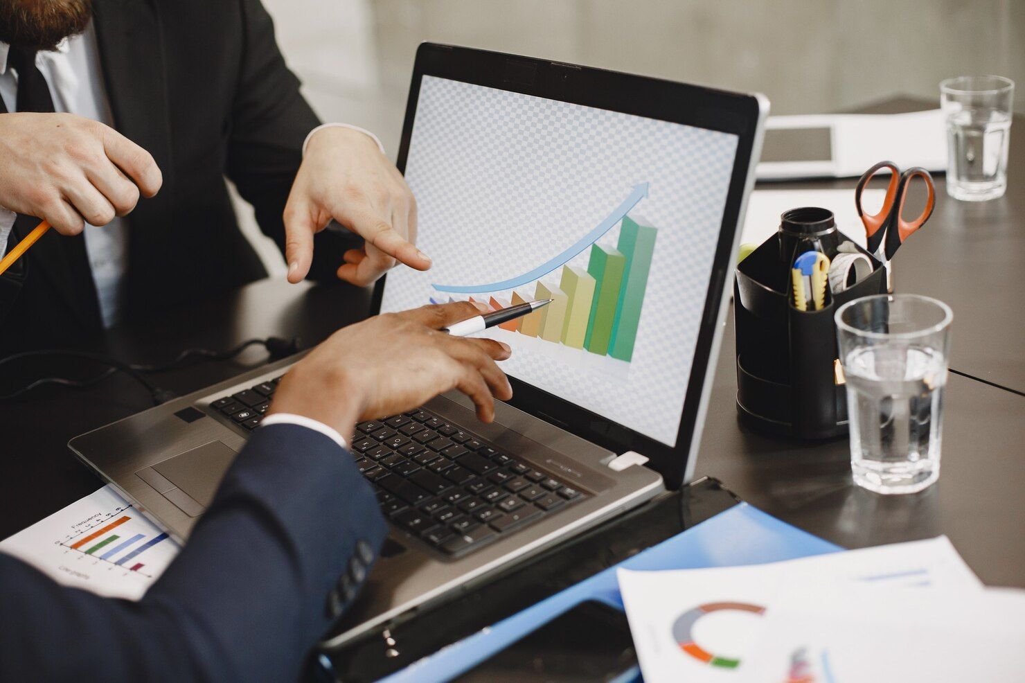 Two people point at a laptop displaying a colorful bar chart in an office setting.