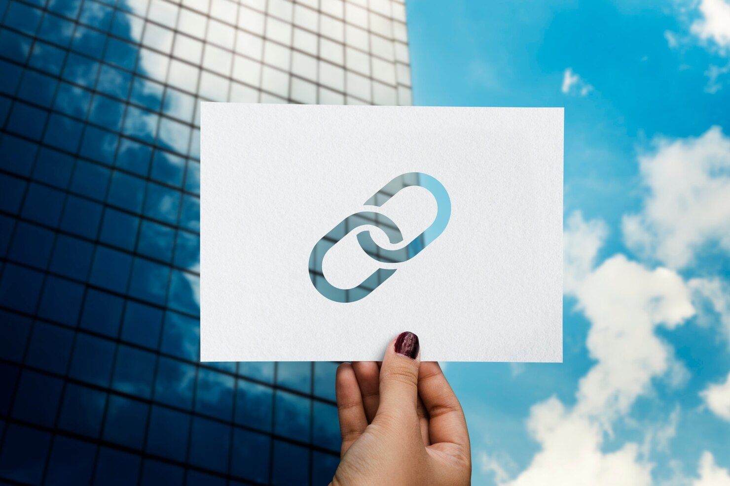 Hand holding a white card with a chain-link icon against a blue sky and glass building background