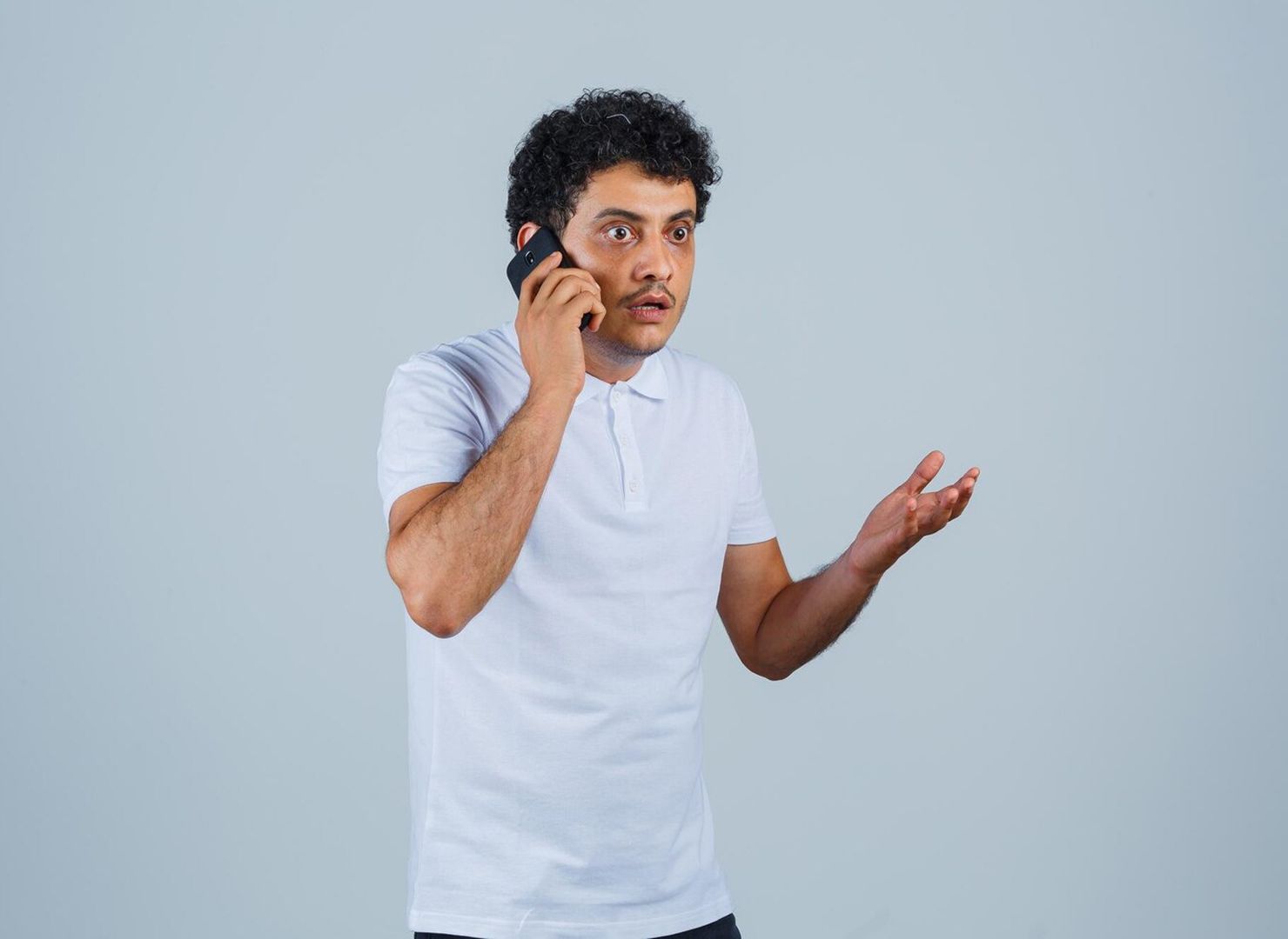 Man in a white shirt talking on a phone, gesturing with one hand against a plain gray background