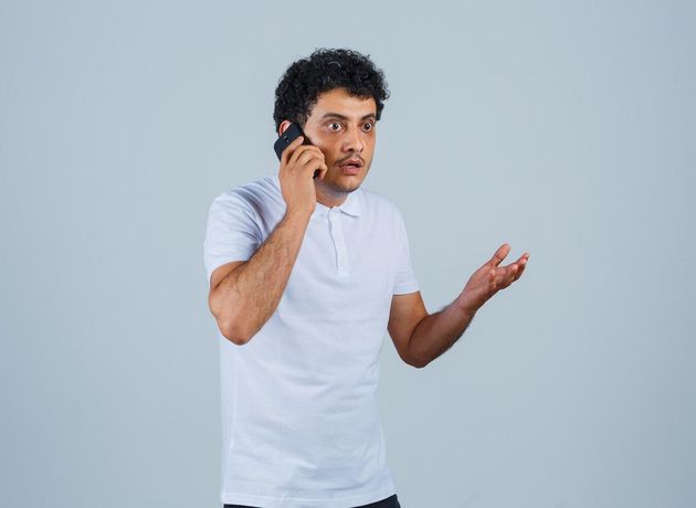 Man in a white shirt talking on a phone, gesturing with one hand against a plain gray background
