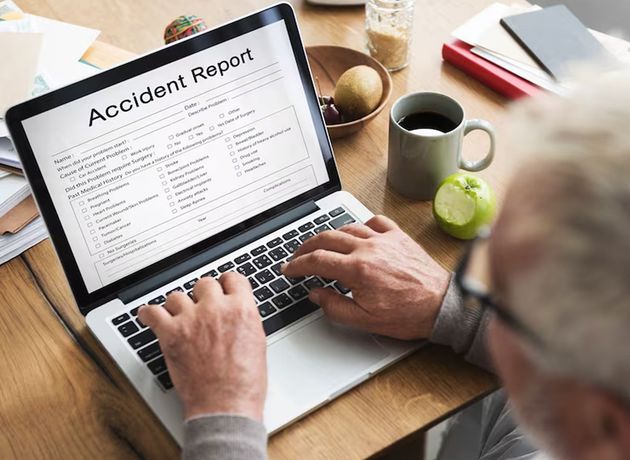 Person typing on a laptop showing an accident report form on a wooden desk with coffee and papers nearby