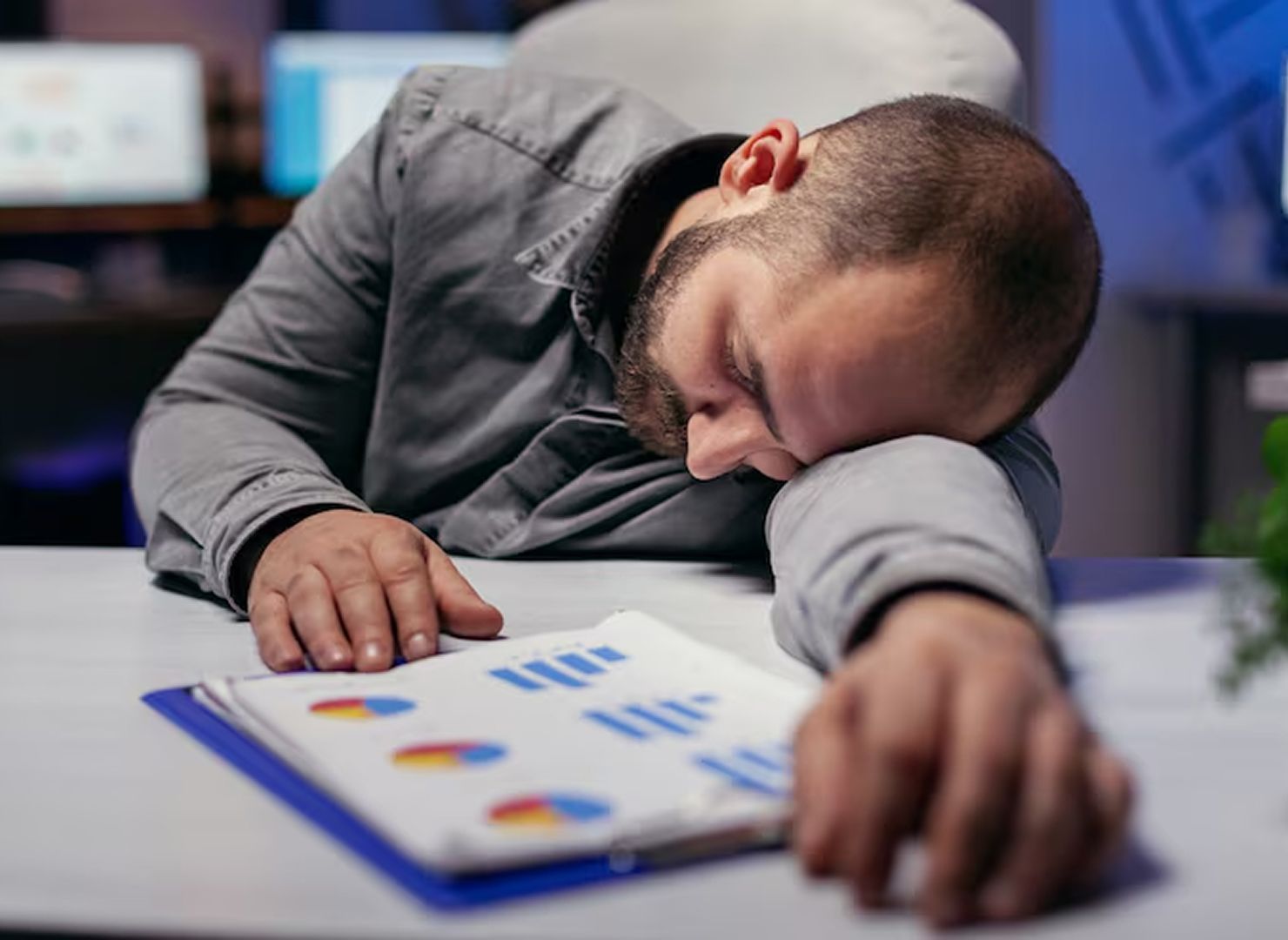 Man sleeping face-down on a desk beside scattered papers and charts