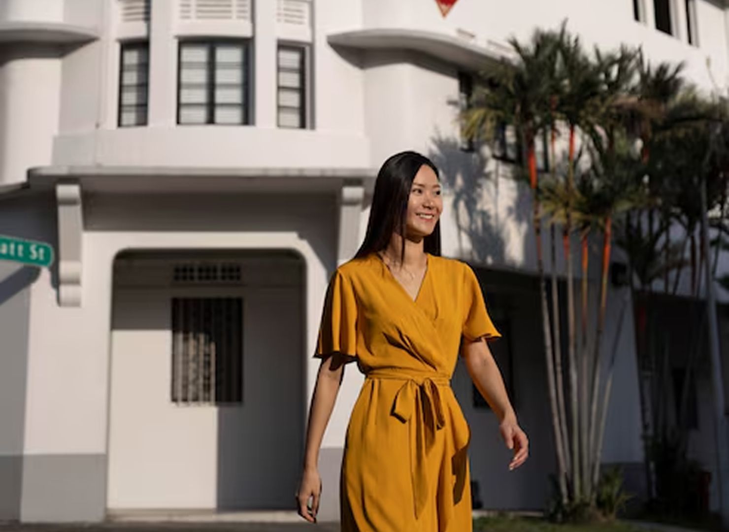 Woman in a mustard yellow dress walking past a white building with palm trees.