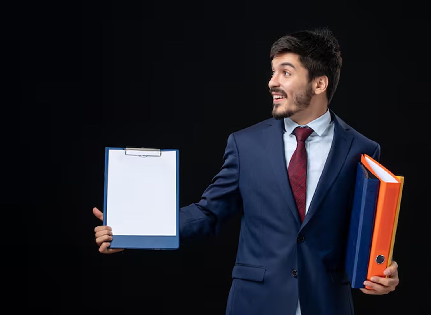 Smiling man in navy suit holding a clipboard and orange binder against a black background