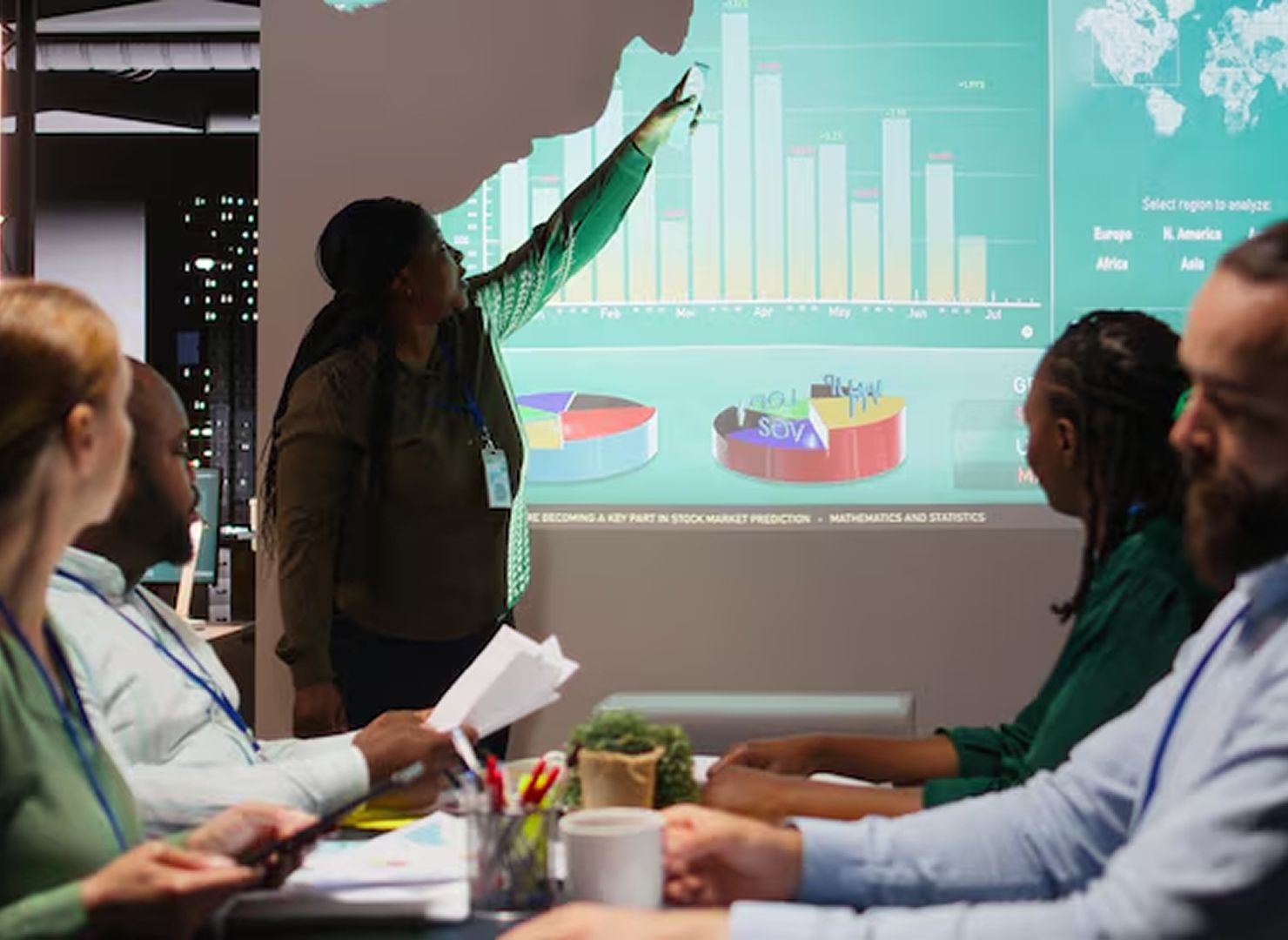 Presenter points at charts on a projected screen during a meeting with seated coworkers.