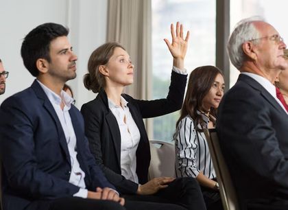 Audience members in business attire listening, one woman raising her hand in a meeting room