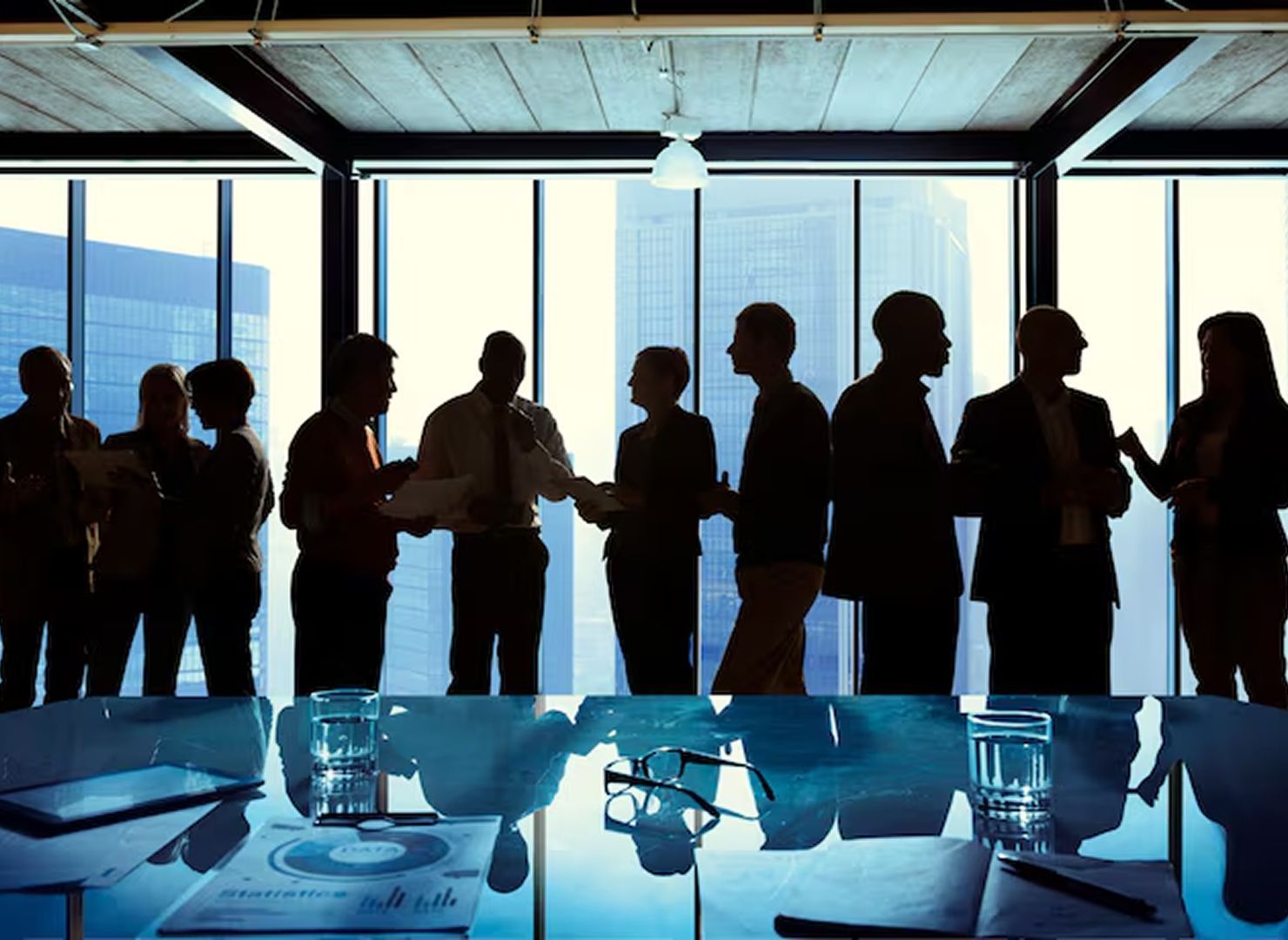 Silhouetted people standing by large windows in a conference room with a table set in the foreground