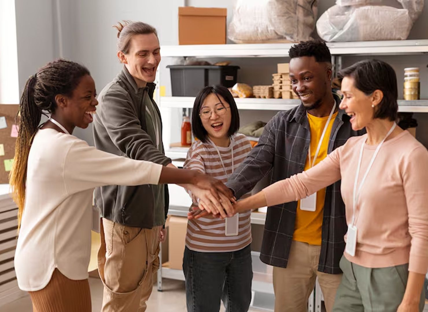 Five people stack hands in a bright room, smiling during a group meeting.