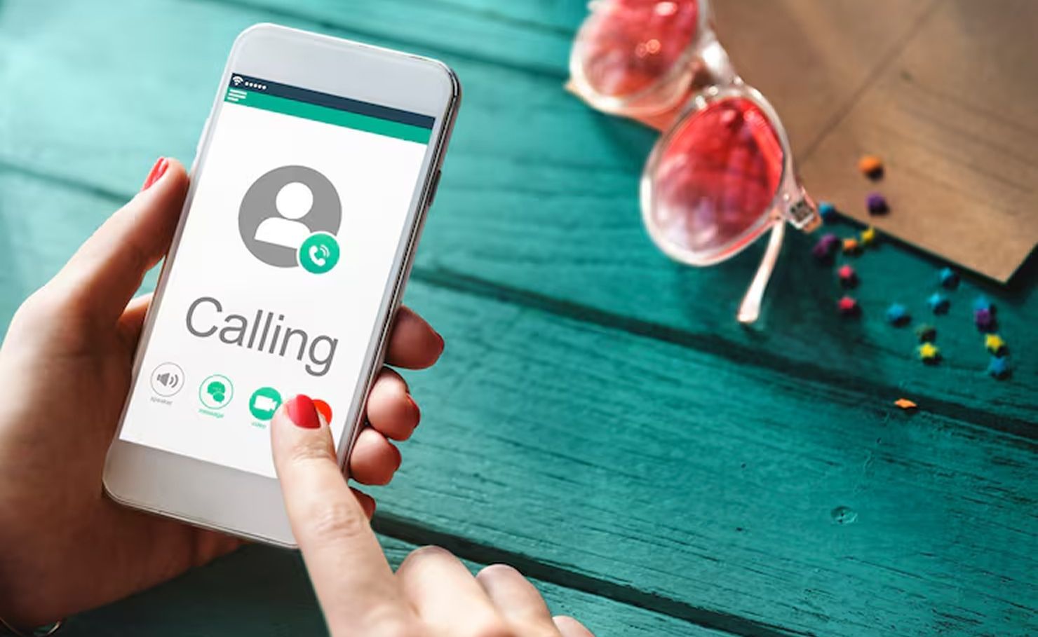 Hand holding smartphone with incoming call screen on turquoise table, with sunglasses and beads nearby