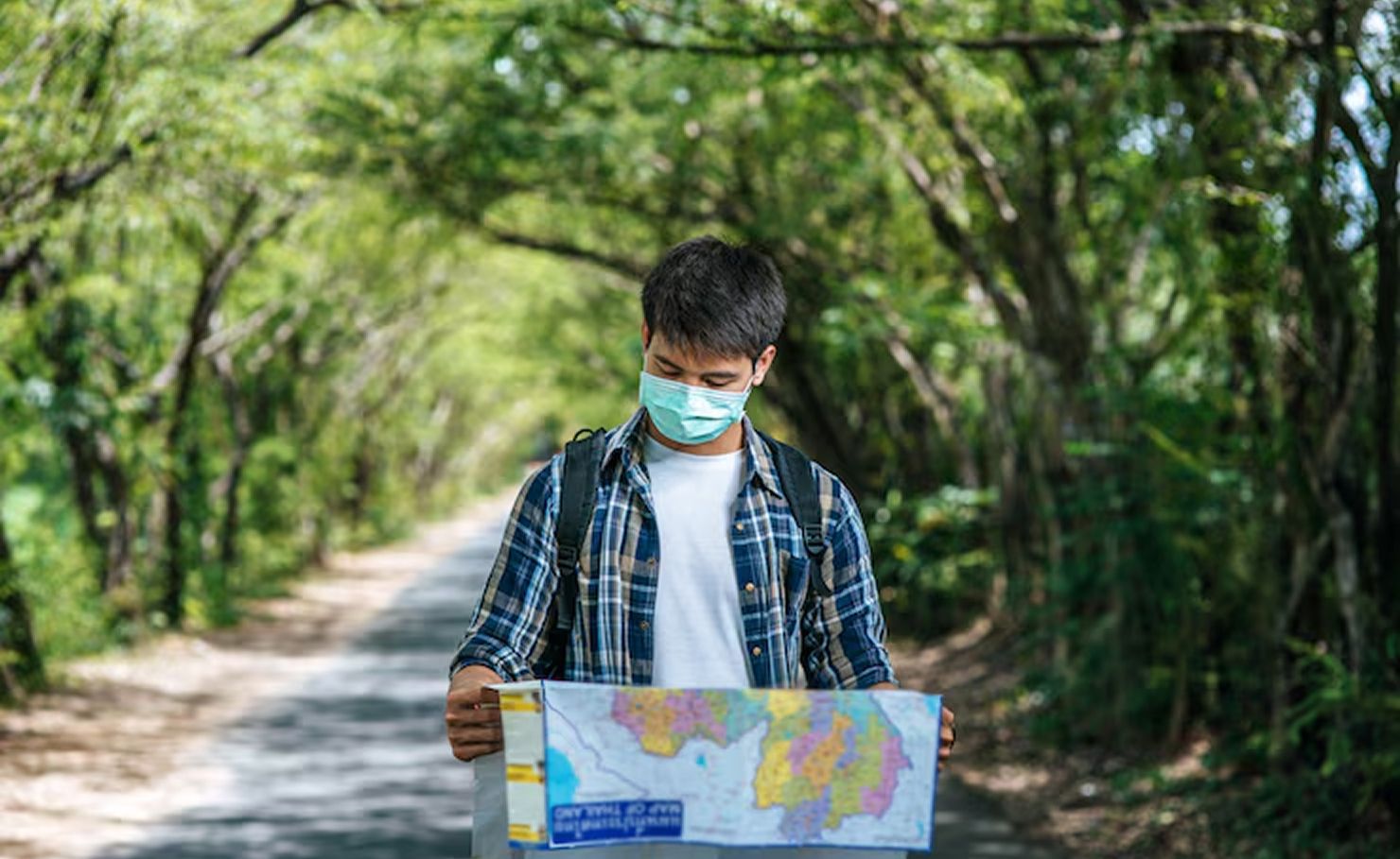 Masked person holding a colorful map on a tree-lined path
