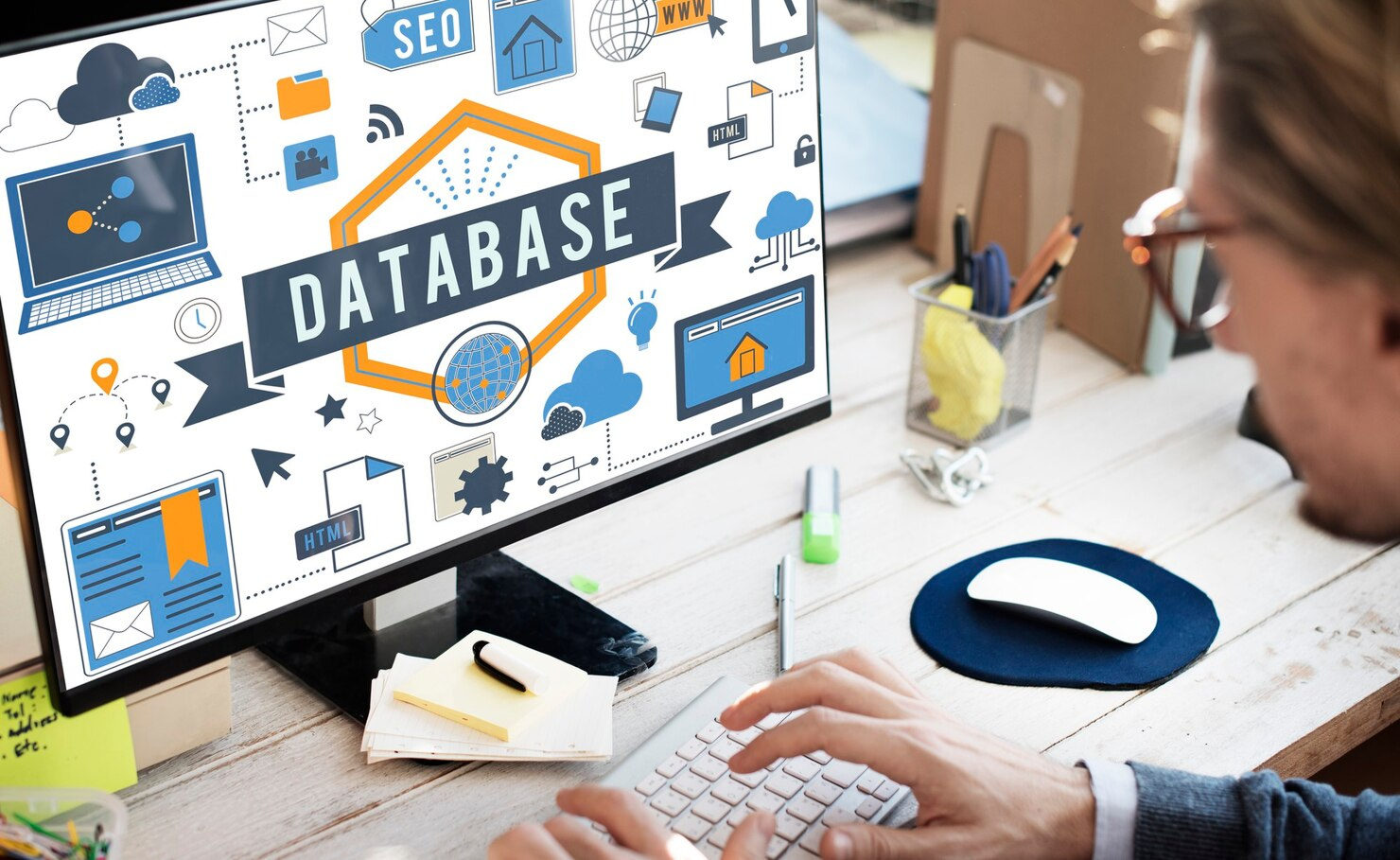 Person typing at a desk beside a monitor showing database icons and the word “DATABASE”