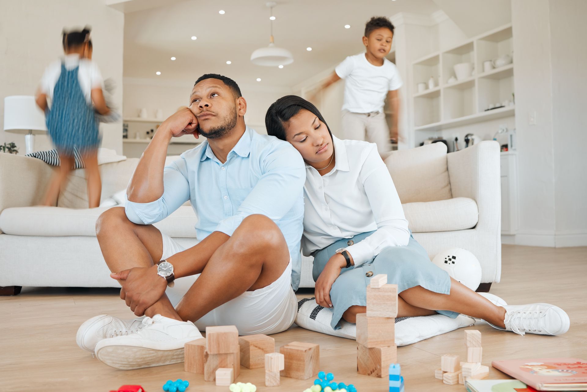 Parents sitting on the floor looking exhausted while children play energetically in the background, illustrating family stress and emotional strain after a traumatic event.