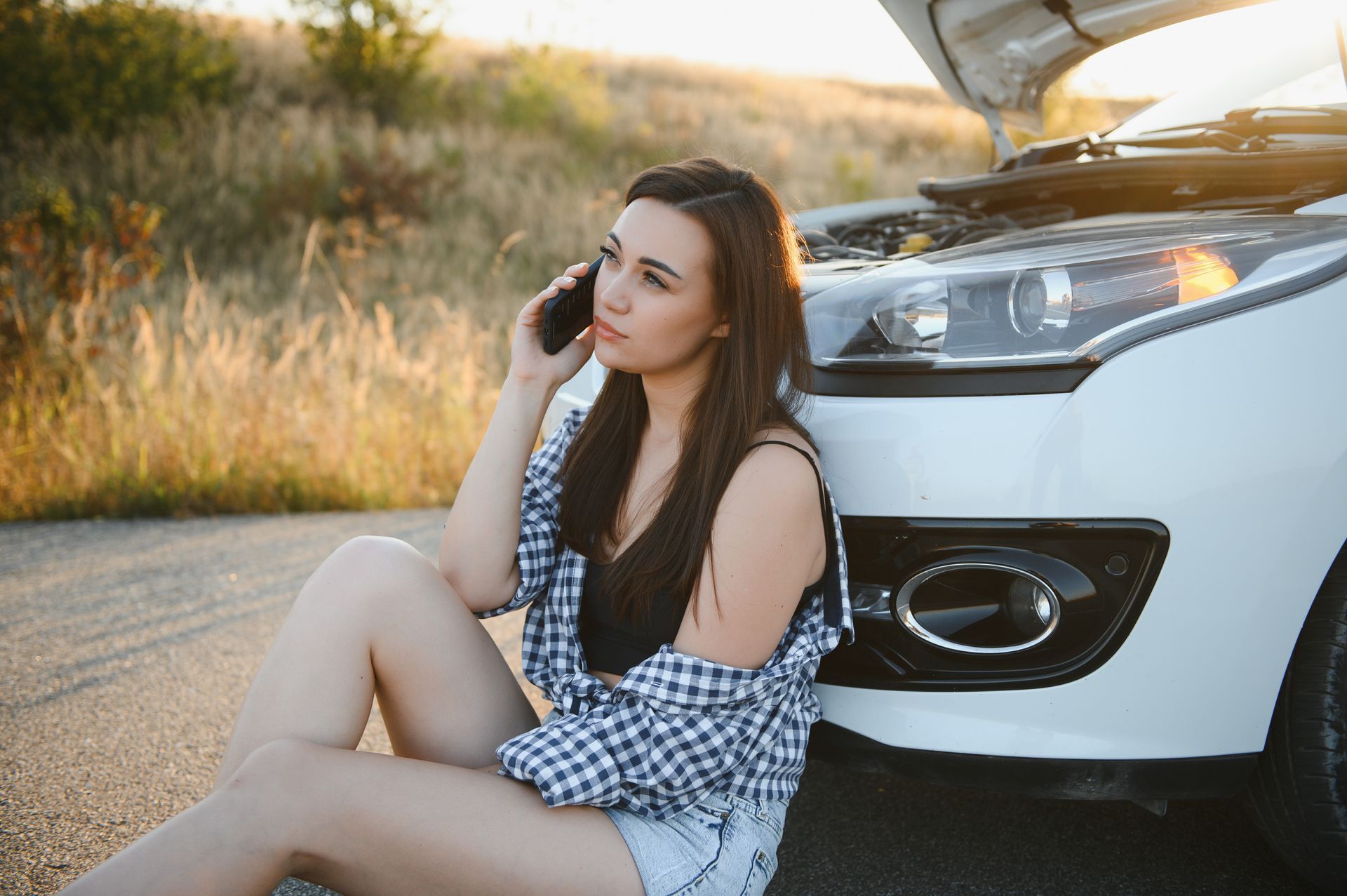 Driver sitting next to a car with the hood open while calling for help after a roadside breakdown