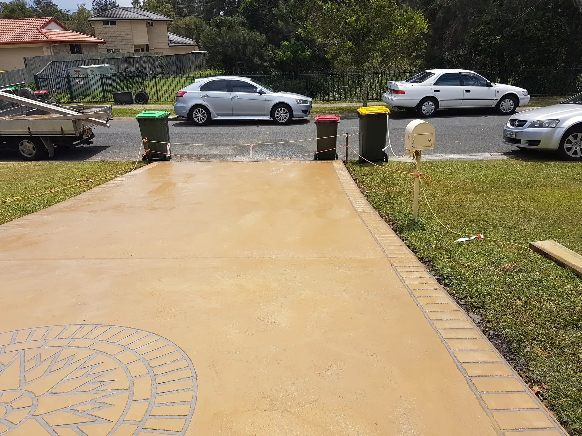 Driveway with three trash bins, mailbox, and parked cars on a suburban street.