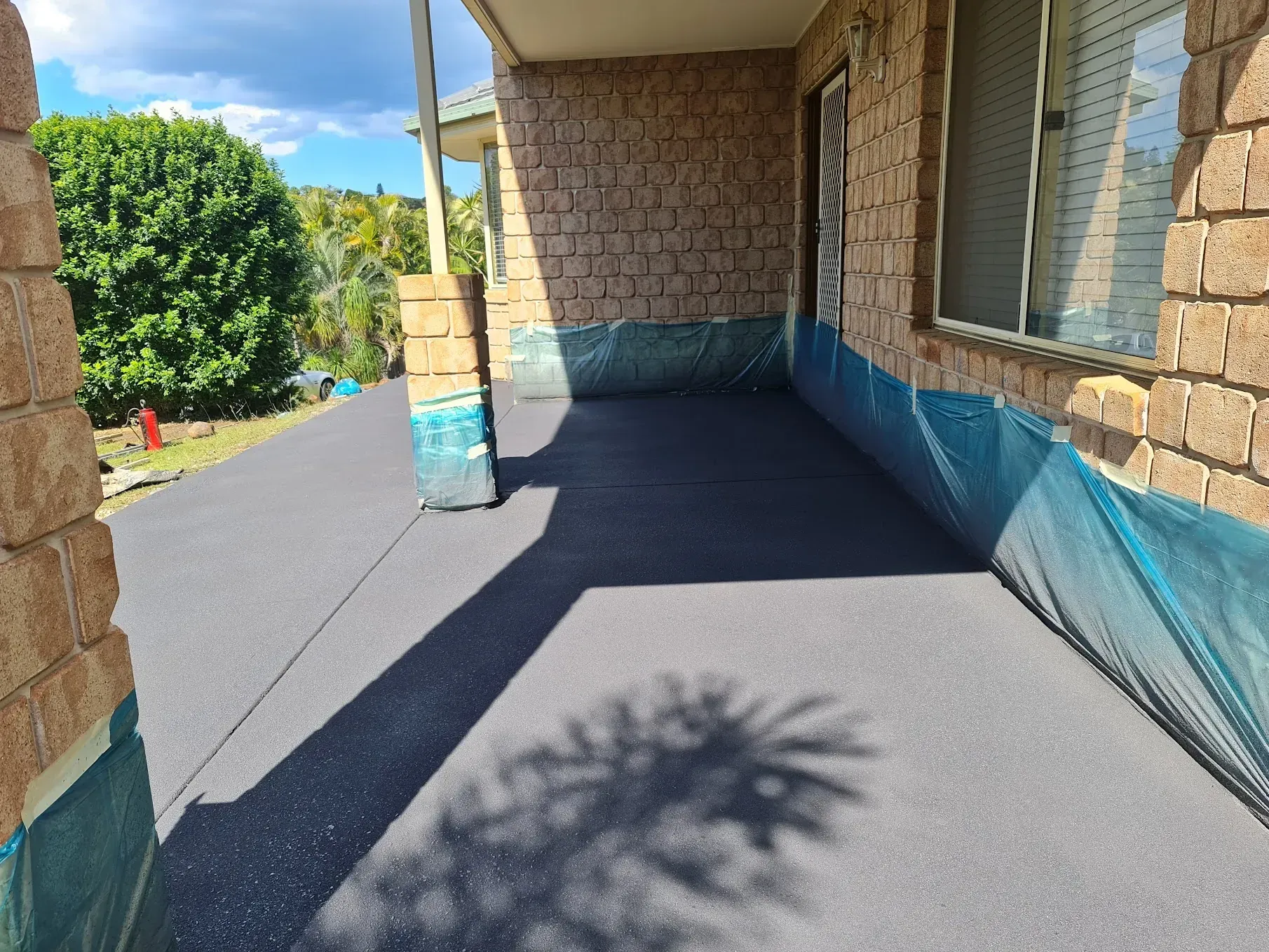 Dark, freshly sealed porch; brick columns and wall; blue netting protects surrounding areas; sunny day.