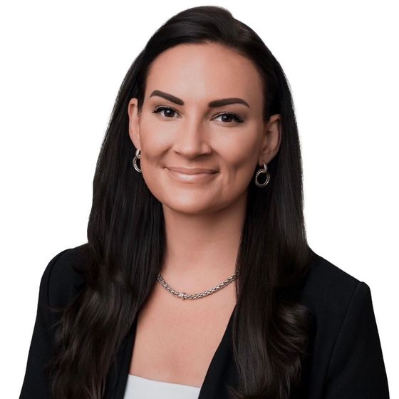 Studio portrait of a woman with dark hair, wearing a black blazer, white top, and silver jewelry, smiling on white background