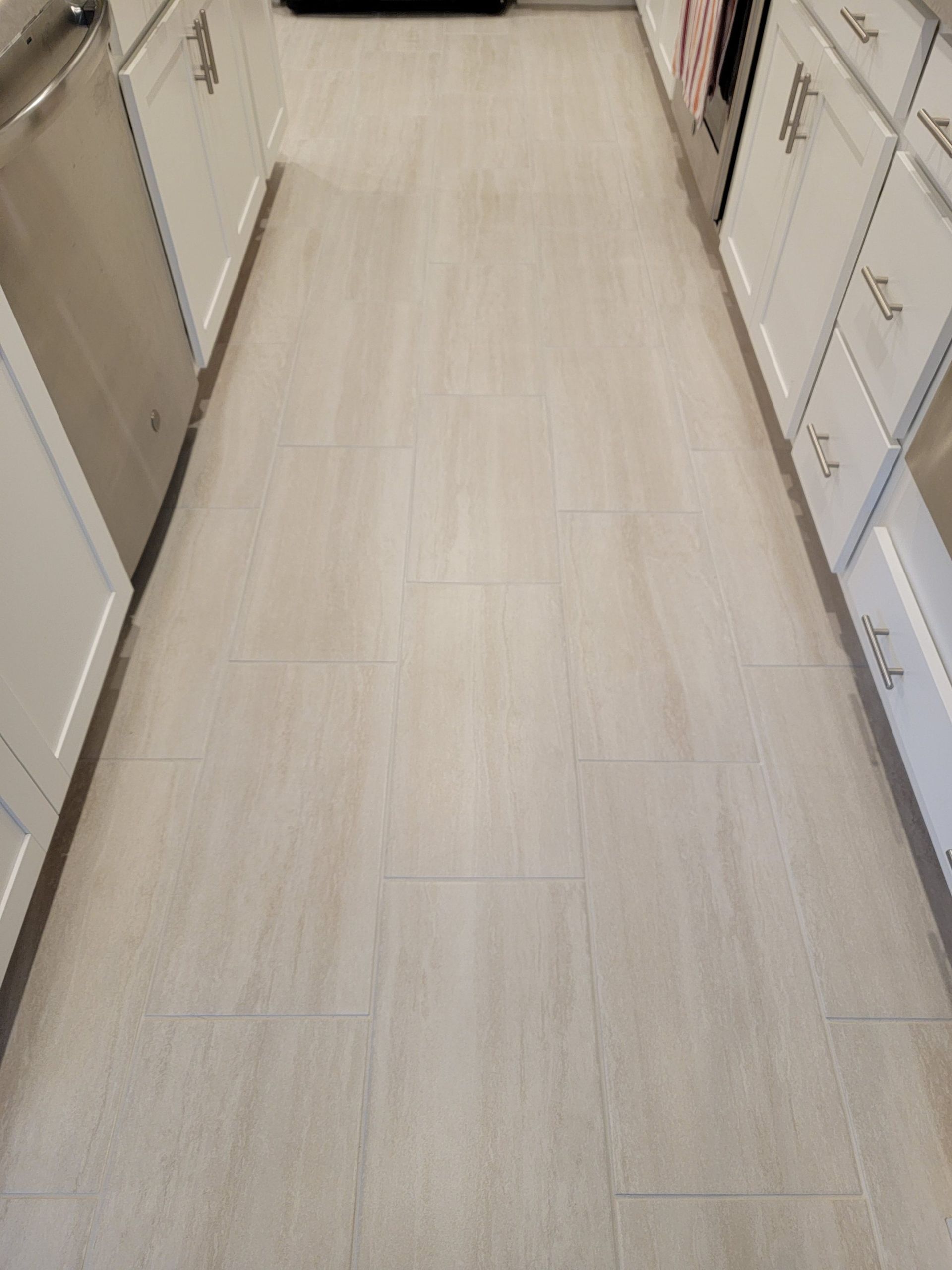 A high-angle view of a narrow kitchen floor with light-colored, wood-look rectangular tiles flanked by white cabinets.