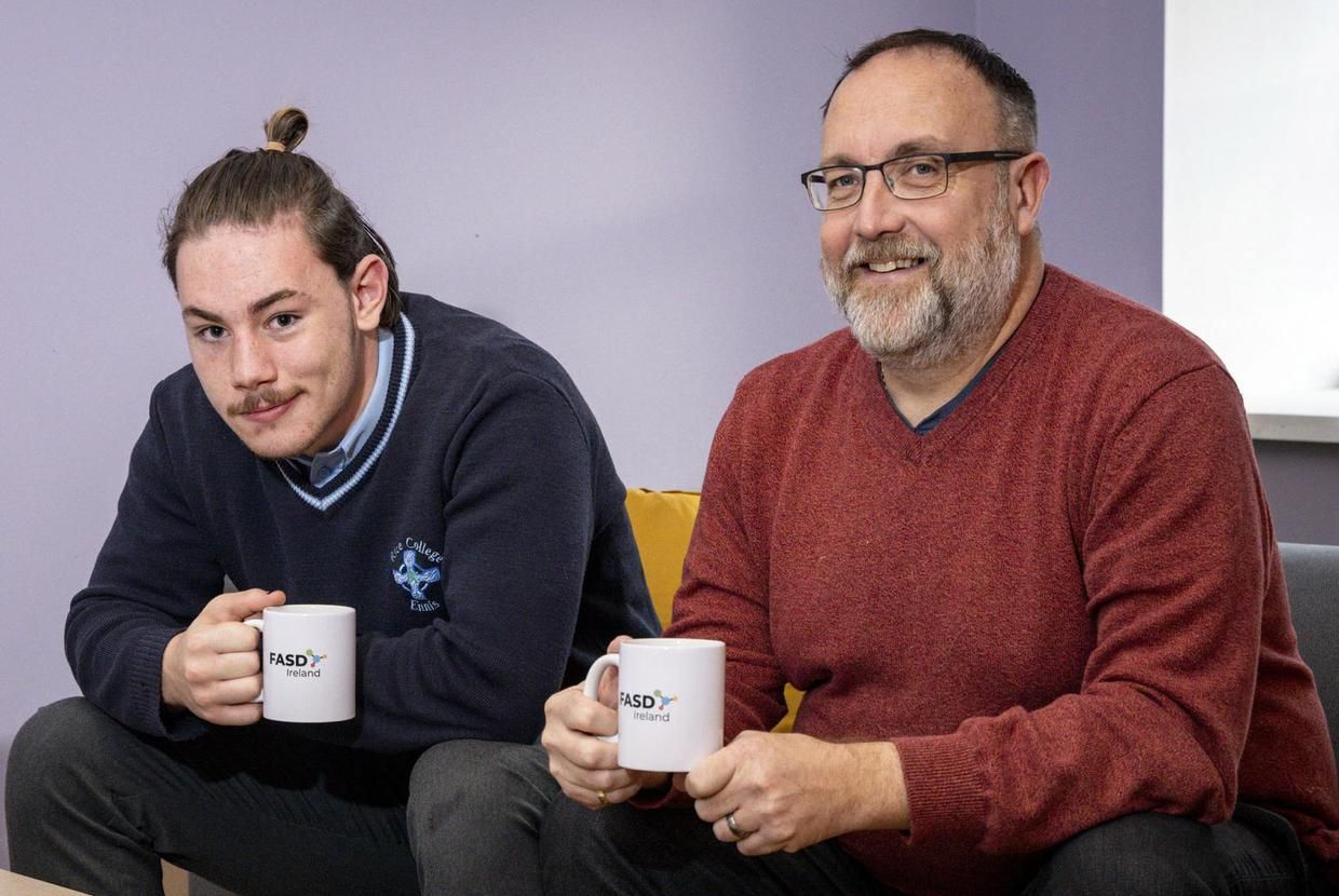 A man and a boy drinking a cup of tea on a sofa looking at the camera