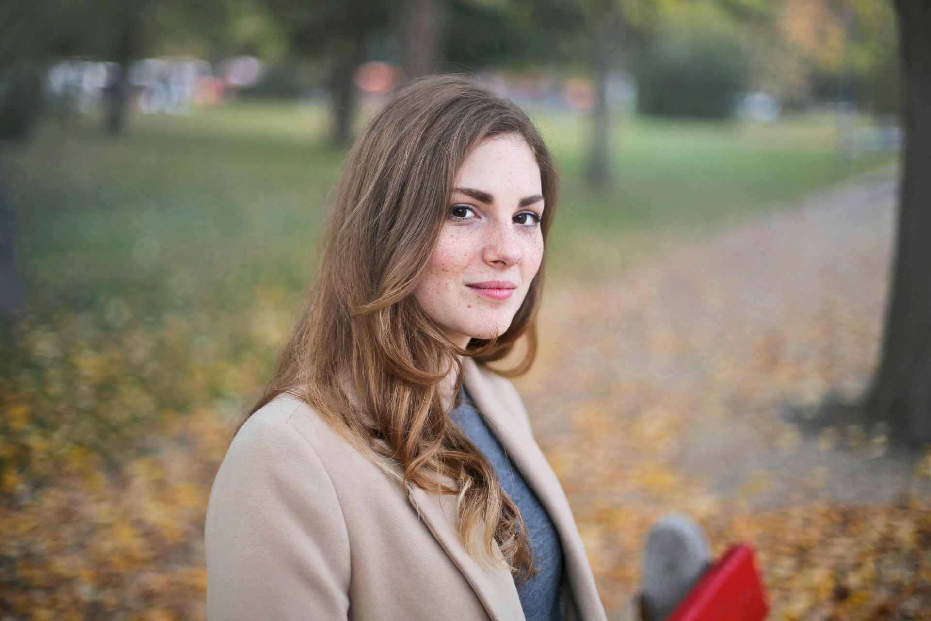 A woman in a coat is standing in a park holding a red book.