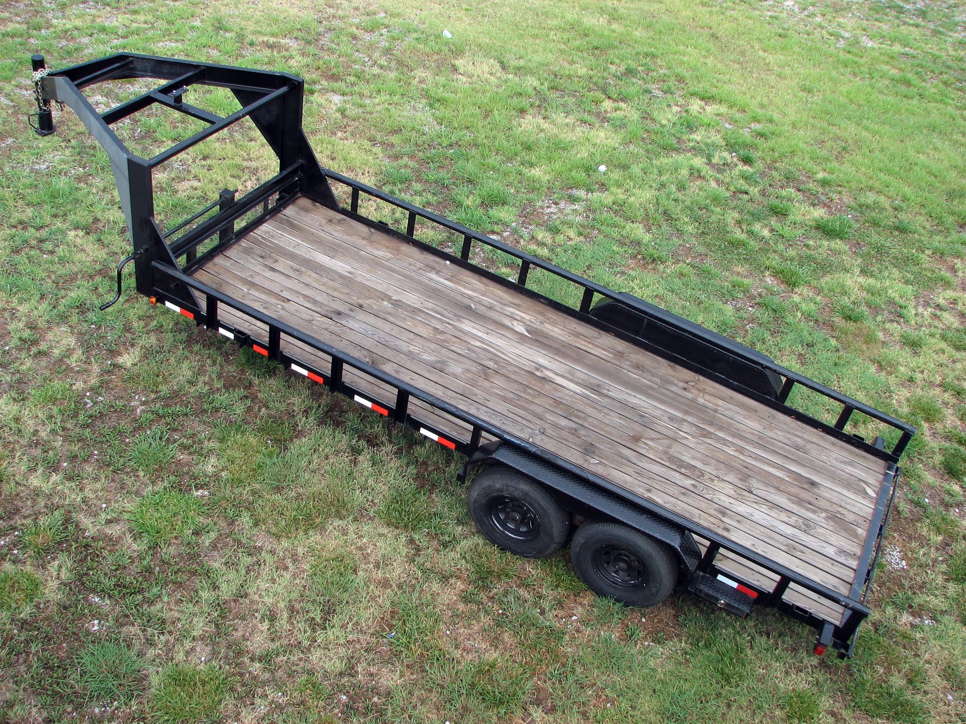Black flatbed utility trailer with wooden deck, two axles, and metal railings on grass.
