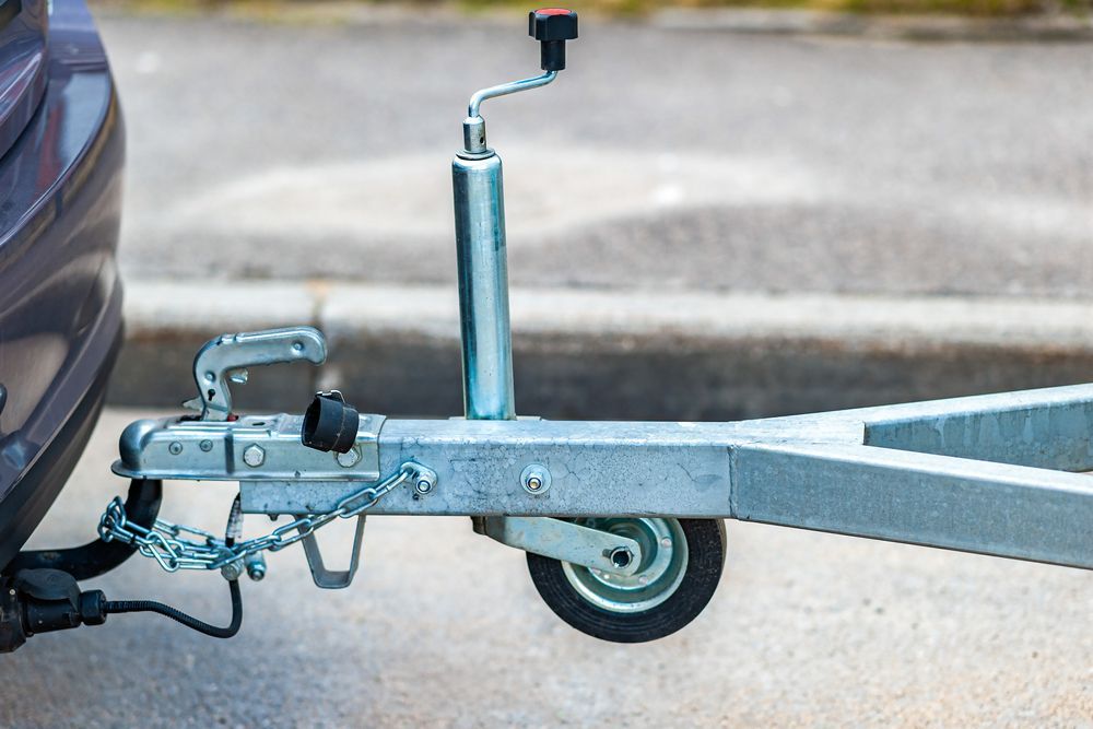 Close-up of a trailer hitch and jack wheel attached to a vehicle in a parking lot
