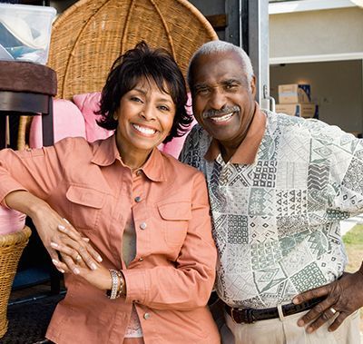 A smiling couple stands in front of a truck filled with moving boxes and furniture, preparing for a move.