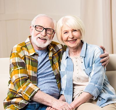 A smiling couple sits closely together on a couch, wearing casual plaid shirts and holding hands.
