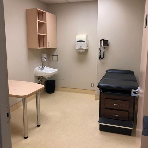 Exam room with sink, exam table, wall dispenser, and beige cabinets in a clinic.