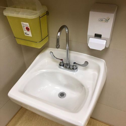 Small beige restroom sink with chrome faucet, soap dispenser, and yellow wall-mounted trash bin.