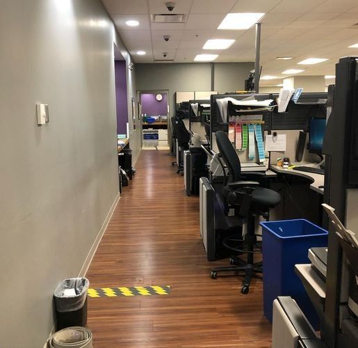 Office hallway with rows of cubicles and desks along a wood floor, viewed down a narrow corridor.