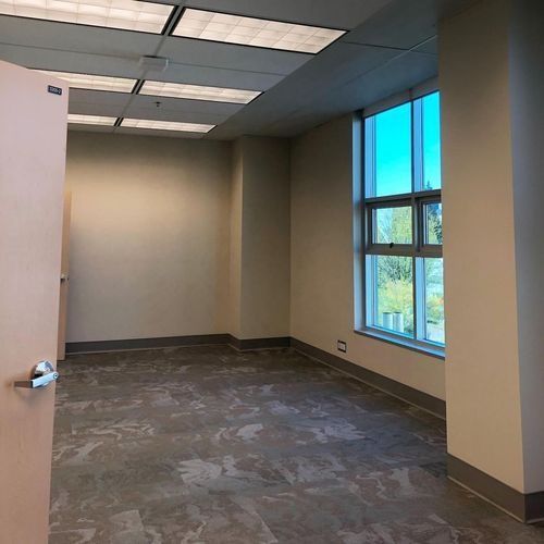 Empty office room with beige walls, patterned carpet, fluorescent ceiling lights, and a large window.