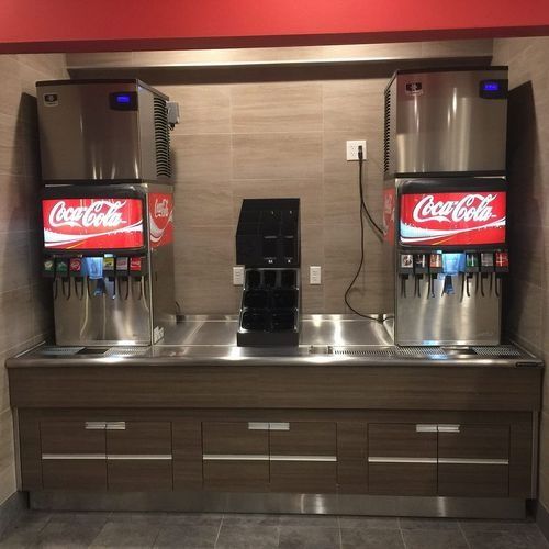 Two Coca-Cola soda fountains on a stainless steel drink station with cabinets and a backsplash