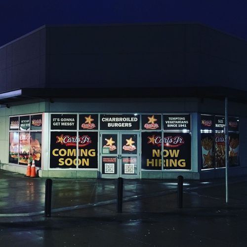 Nighttime storefront with neon signs reading “Coming Soon” and “Now Hiring” on a wet street corner.
