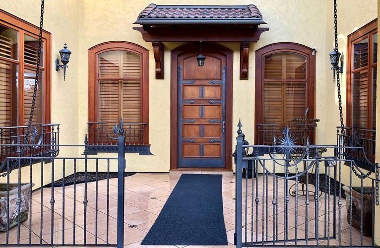 Front entrance of a yellow house with a brown wooden door, shuttered windows, and black iron gate.