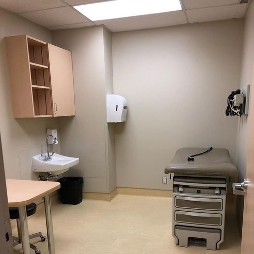 Empty medical exam room with exam table, sink, cabinets, and wall-mounted medical equipment