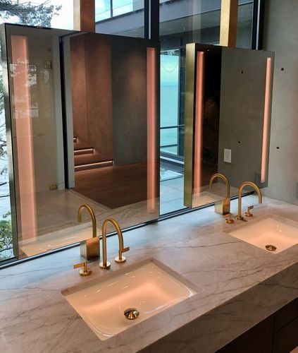 Modern restroom vanity with two sinks, brass faucets, and large mirrors reflected in glass panels.