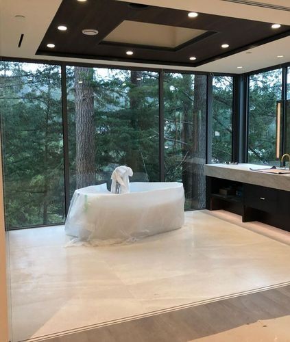 Modern living room with floor-to-ceiling forest views, black ceiling lights, and a white chair wrapped in plastic.