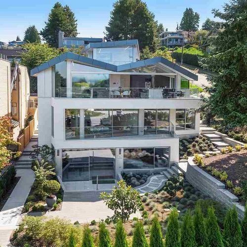 Modern multi-story white house with large glass windows and terraced landscaping on a hillside