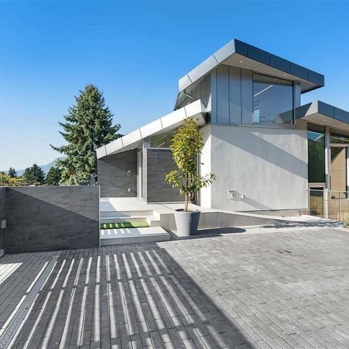 Modern gray house with large glass windows, flat roof, and a striped driveway under bright sunlight